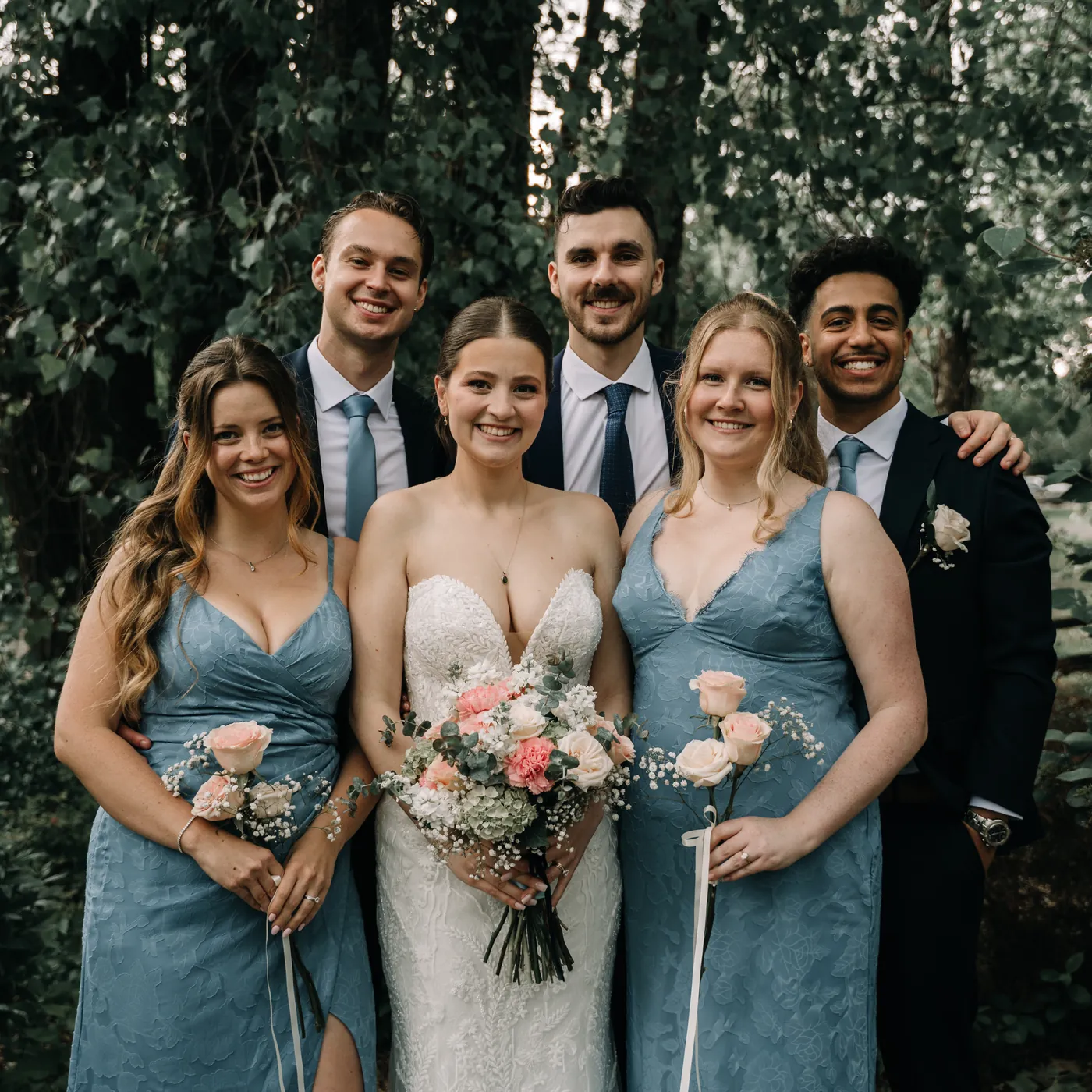 The wedding party with the bride and her bouquet, bridesmaids in blue and the groomsmen, against a leafy green backdrop.